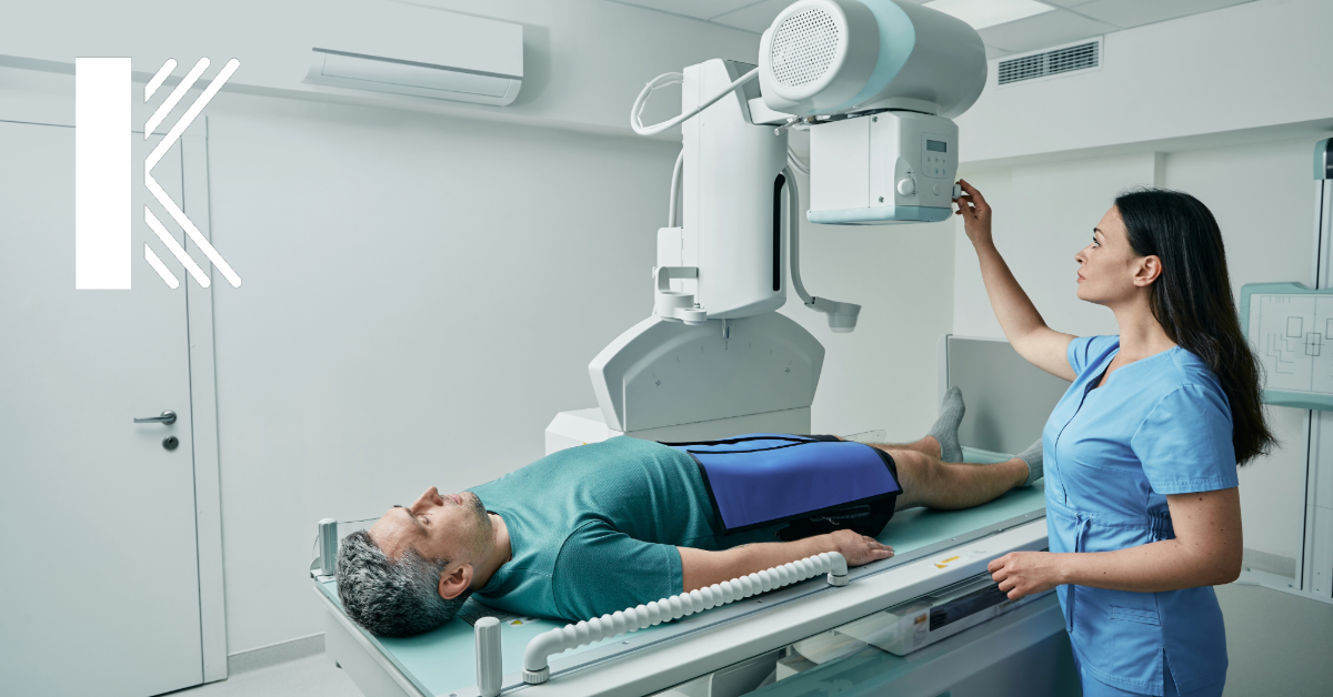 The image shows a healthcare professional in a clinical setting operating an X-ray machine. A patient lies on the examination table, ready for the procedure. The room is clean and modern, with the Kastner Insurance Group logo subtly placed in the corner. The image highlights the importance of protecting expensive medical equipment like X-ray machines, which are critical in medical practices.