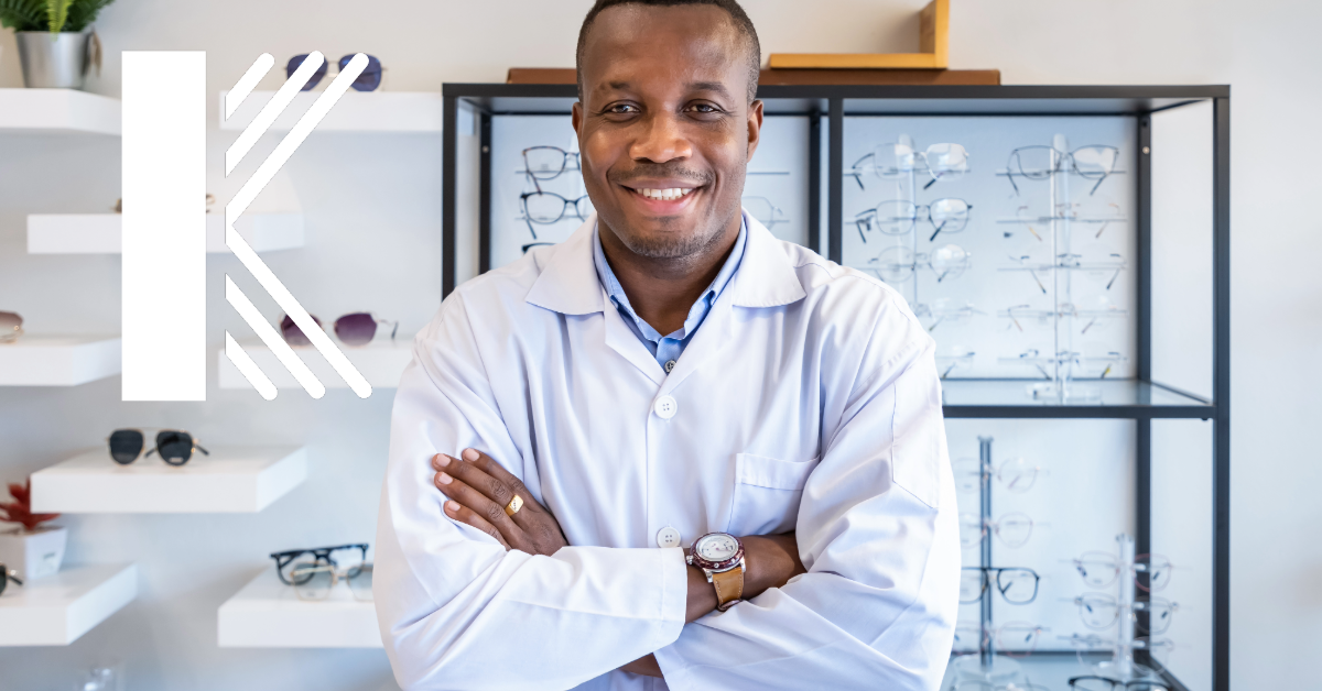 A confident optometrist in a white lab coat stands with arms crossed in front of a display of eyeglasses. The large “K” logo is displayed on the left side of the image.