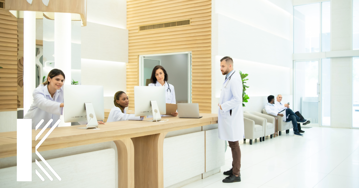 A modern medical office reception area with a sleek, wooden front desk. Four healthcare professionals in white lab coats are engaged in various tasks at the desk—two working on desktop computers, one using a laptop, and one standing and conversing. In the background, two more healthcare professionals are seated and chatting. The environment is clean and well-lit with light wood and white decor, creating a welcoming and professional atmosphere. The Kastner 