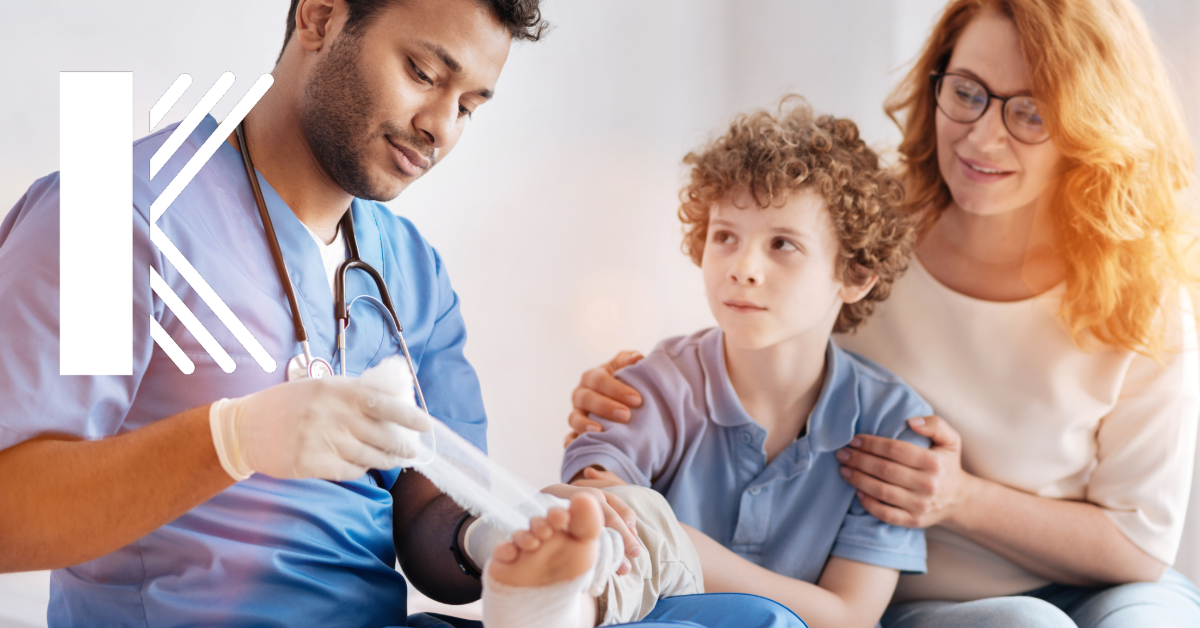 A healthcare provider in a blue uniform is wrapping a young boy’s foot while his mother sits beside him, offering support. The healthcare professional is focused on the child, who looks up at him while receiving care at an urgent care center.